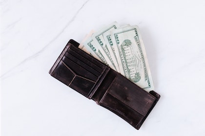 From above of black leather wallet with pockets filled with heap of dollar banknotes on white table